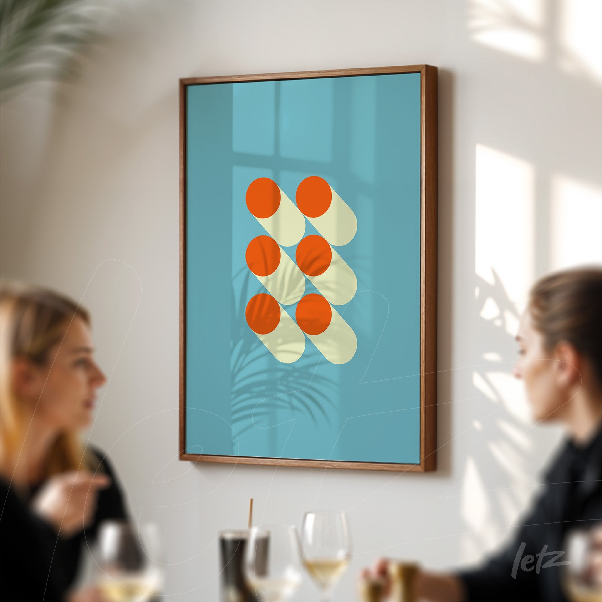 framed geometric art with orange circles and pale yellow shadows on a blue background, set in a dark wooden frame, displayed on a white wall with two people nearby