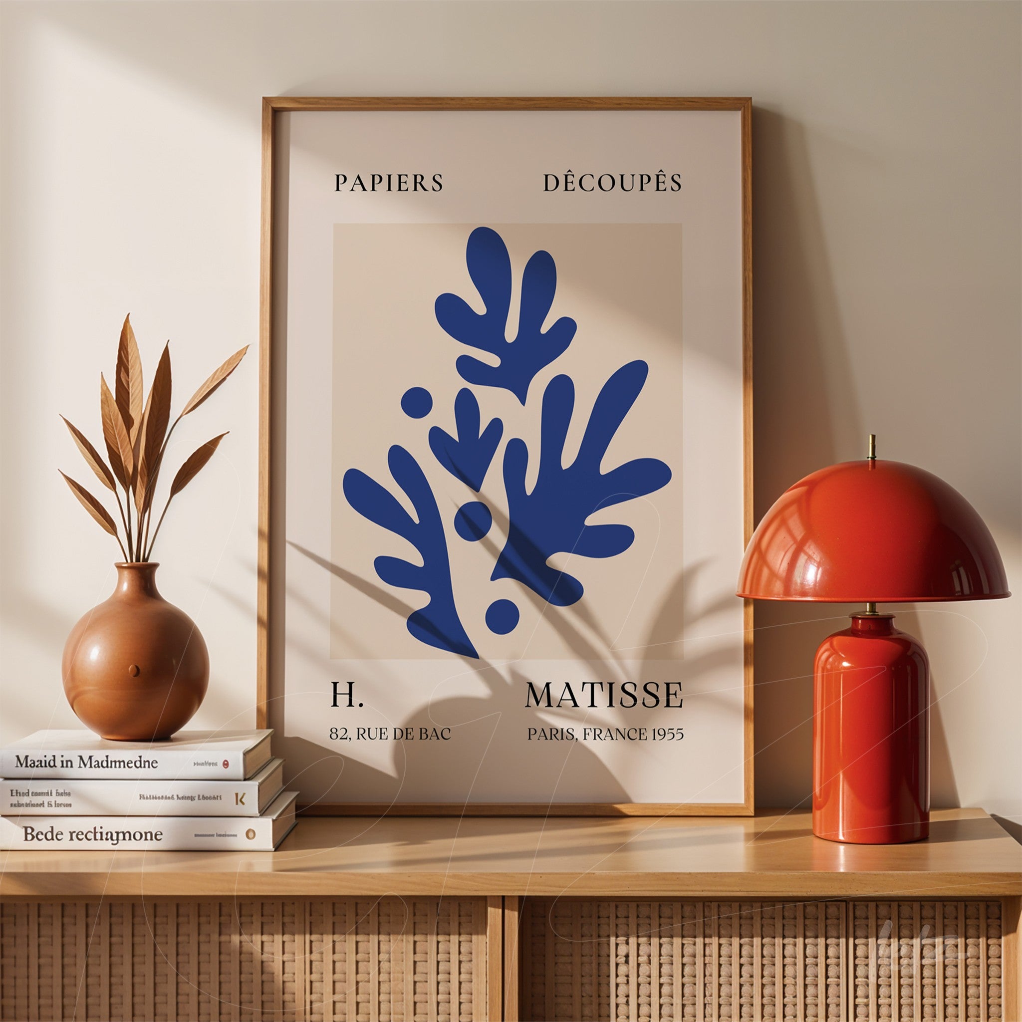 framed abstract wall art in blue depicting organic shapes against a beige background displayed on a wooden shelf beside a red lamp and a vase