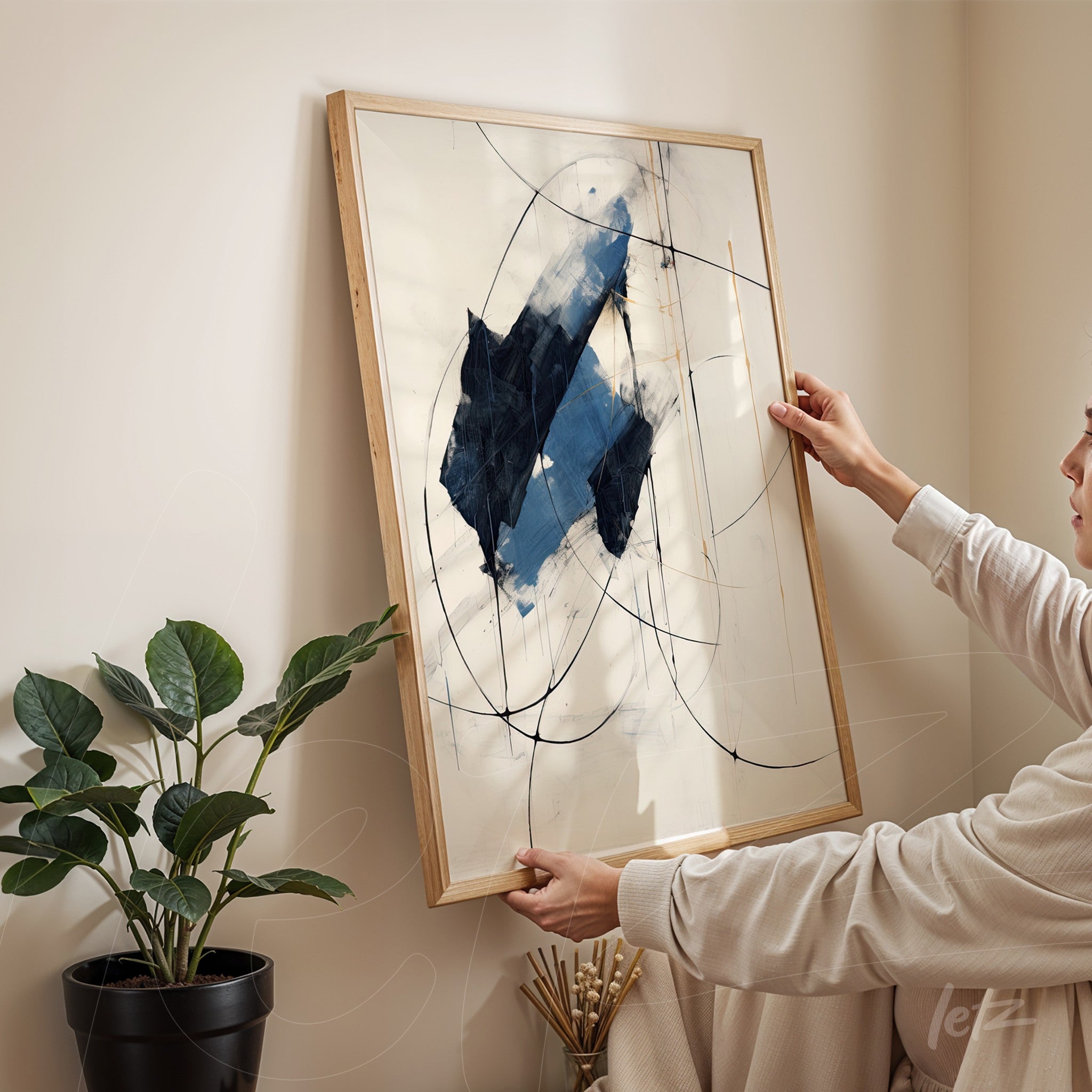 abstract wall art featuring blue elements and black lines, framed in light wood, being held by a person in a minimalist setting with a plant in the background