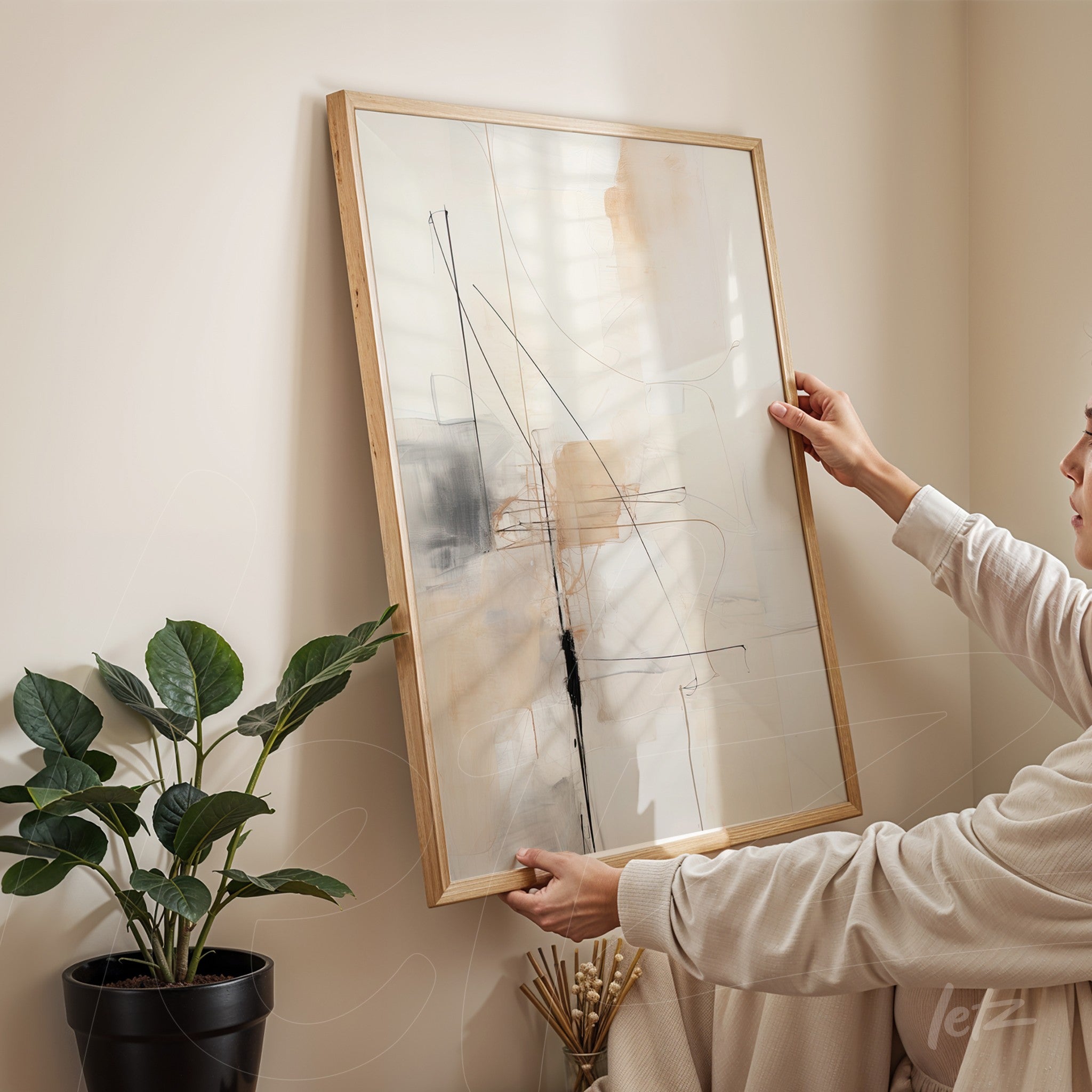 abstract wall art in light wood frame featuring subtle lines and shapes in neutral tones held by a person next to a green plant