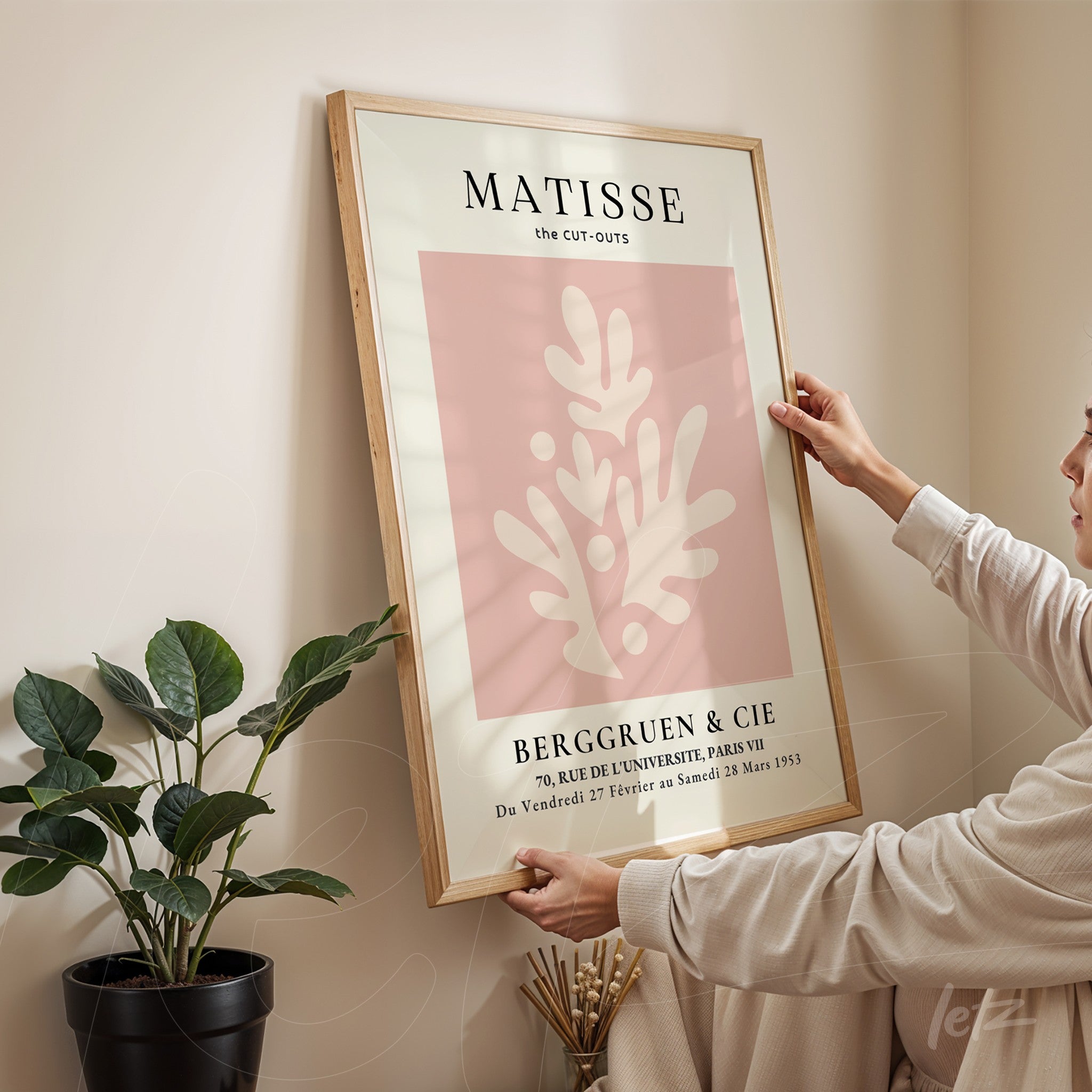 framed modern art print featuring soft-colored foliage design, supported against a wall with a woman adjusting the frame position