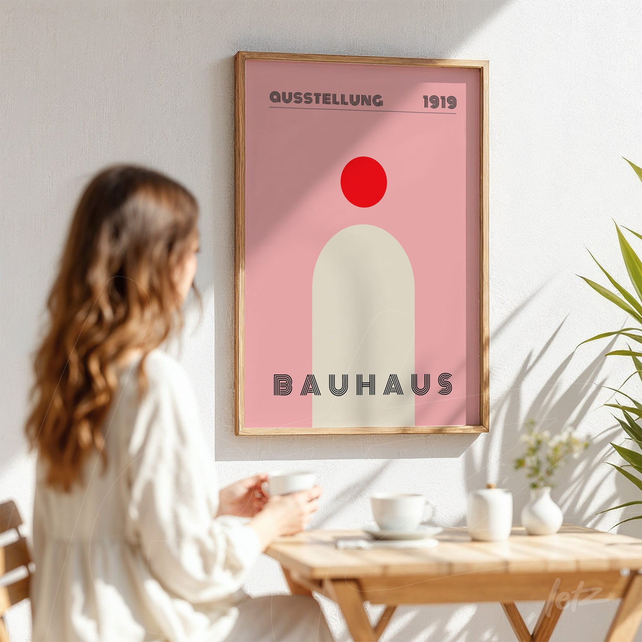 bauhaus style poster print with geometric shapes in pink and red tones, framed in light wood, displayed on white wall next to seated woman