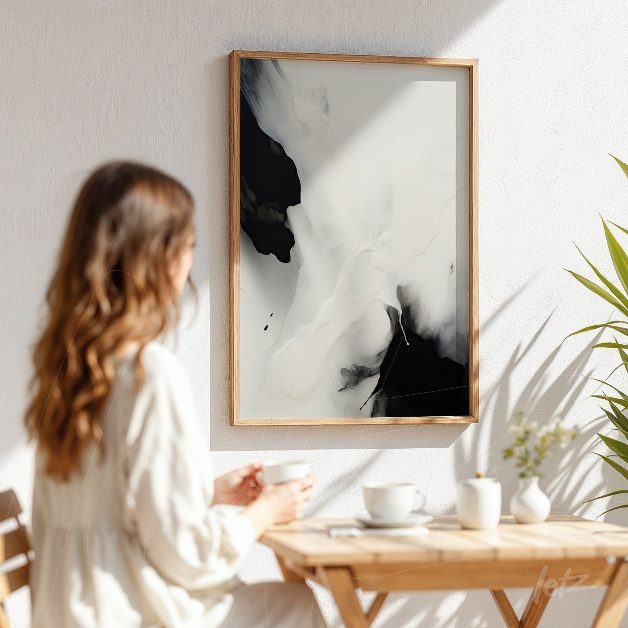 abstract art piece in black and white framed in light wood displayed on a white wall next to a small plant