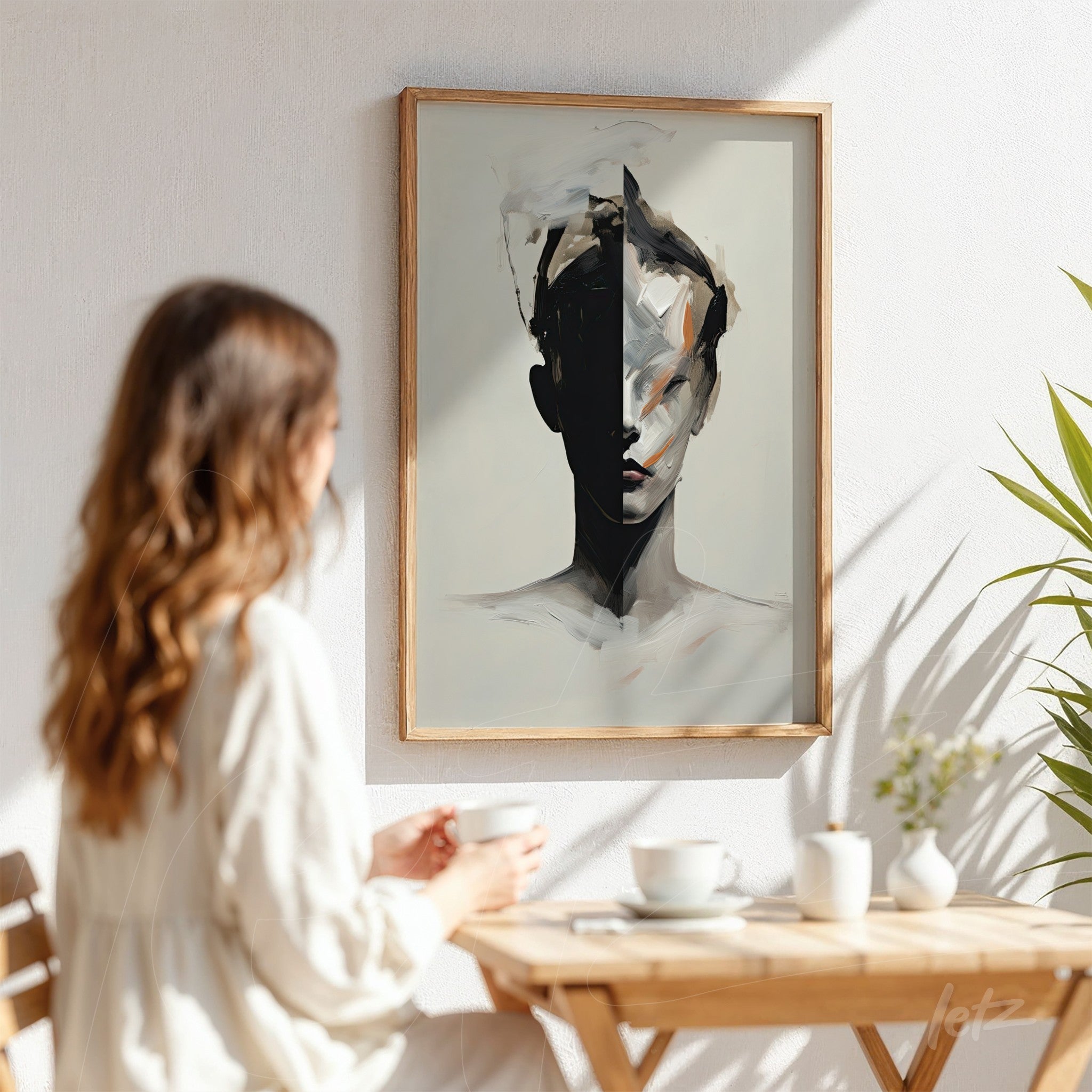 abstract art piece of a partially obscured human face in a light wood frame displayed on a white wall next to a woman sitting with a cup