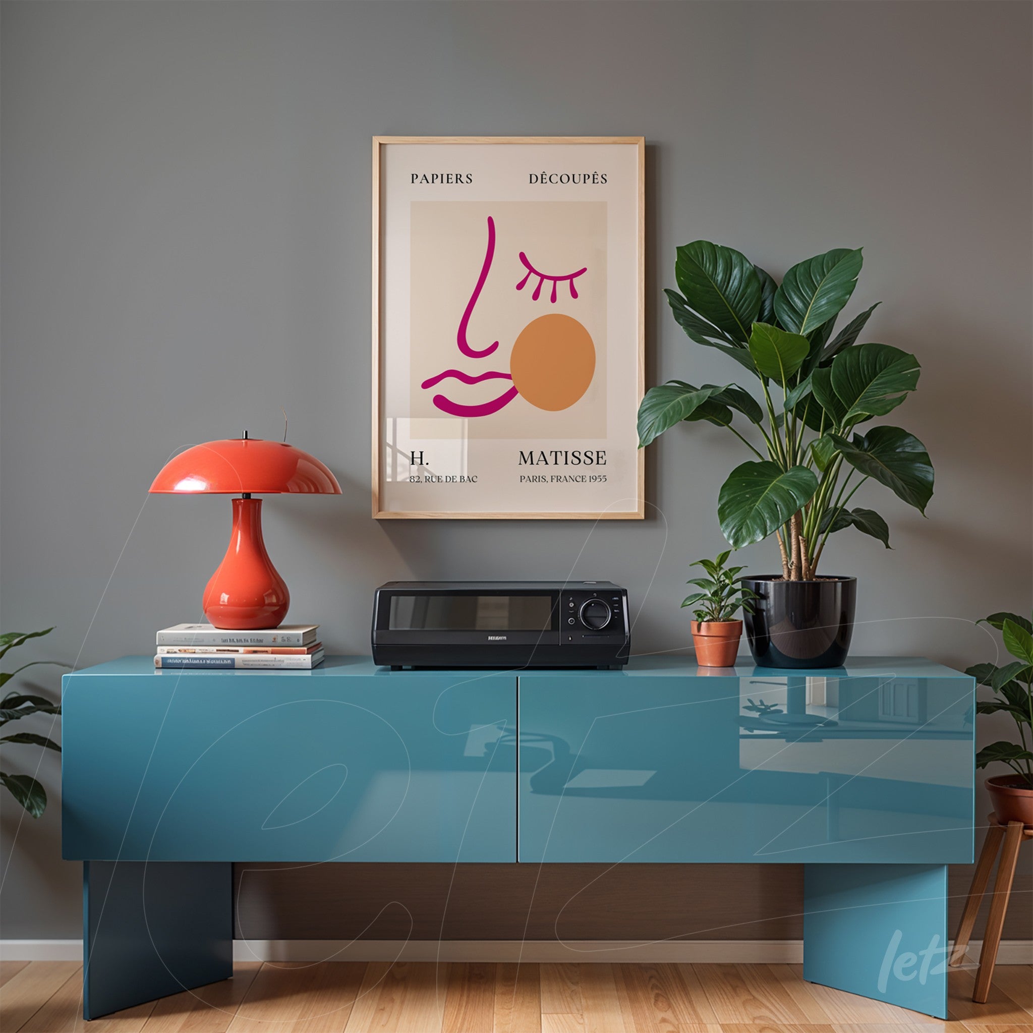 framed modern art print featuring a stylized face in gold frame above a blue table, accompanied by a red lamp and plant