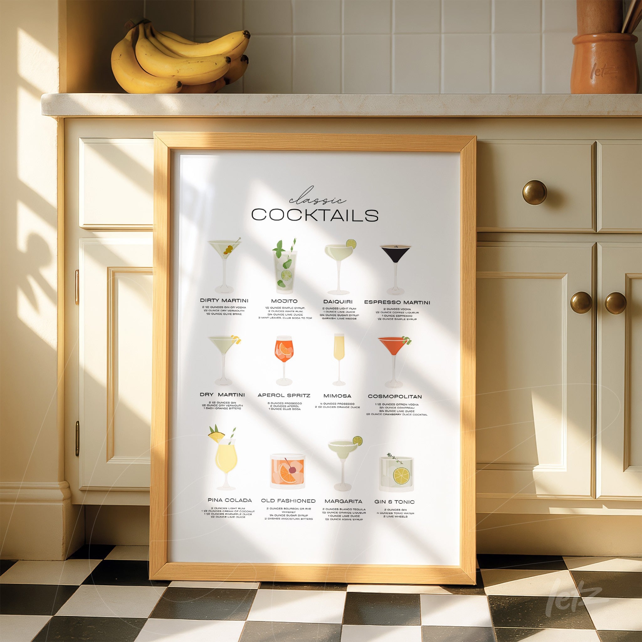 framed art print featuring illustrations of classic cocktails in light wood frame displayed on a kitchen counter with bananas in the background