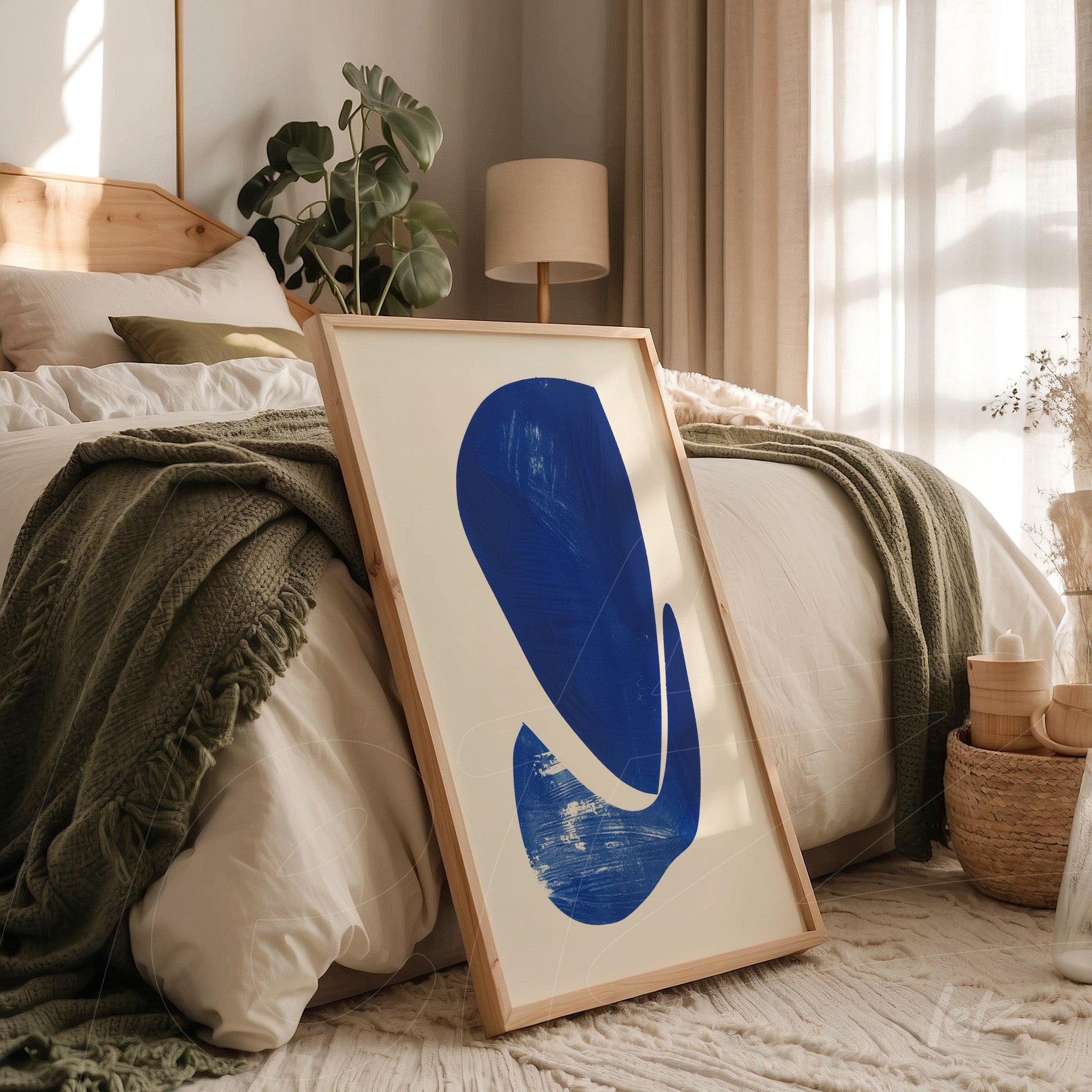 modern artwork featuring an abstract blue shape in a light wood frame leaning against a bed with white sheets and a green throw blanket