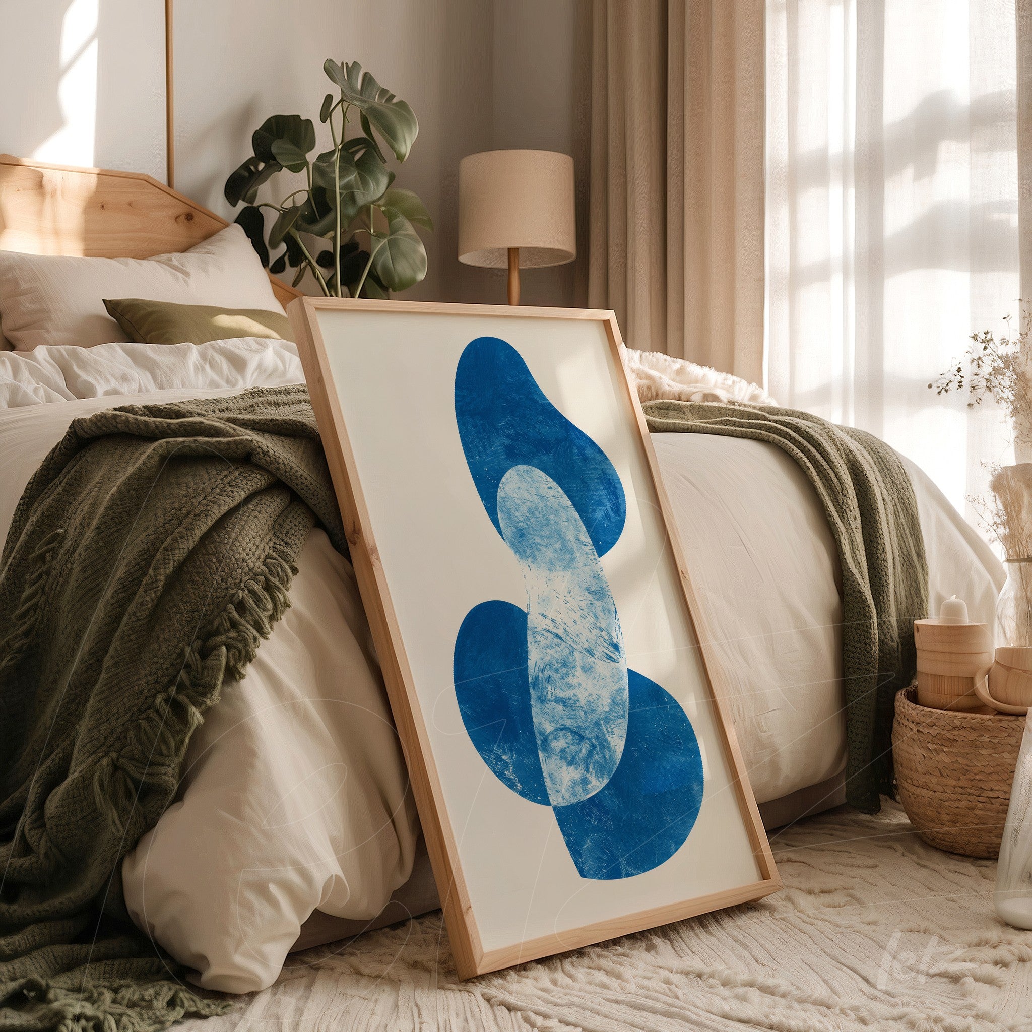 framed abstract art in shades of blue with light wooden frame leaning beside a bed with white bedding and green throw blanket