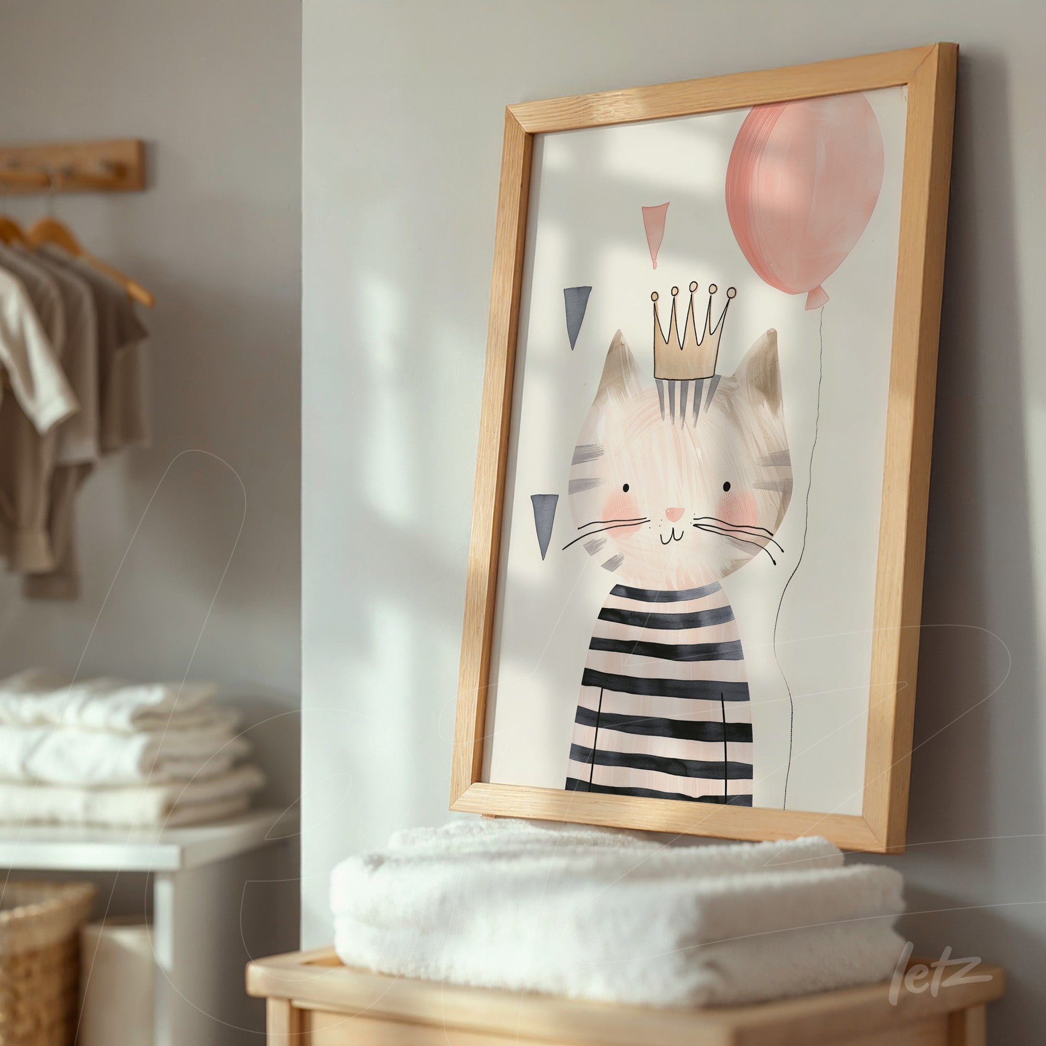 framed decorative art featuring a cat illustration wearing a crown, dressed in stripes and holding a pink balloon, displayed on a stand in a room with towels