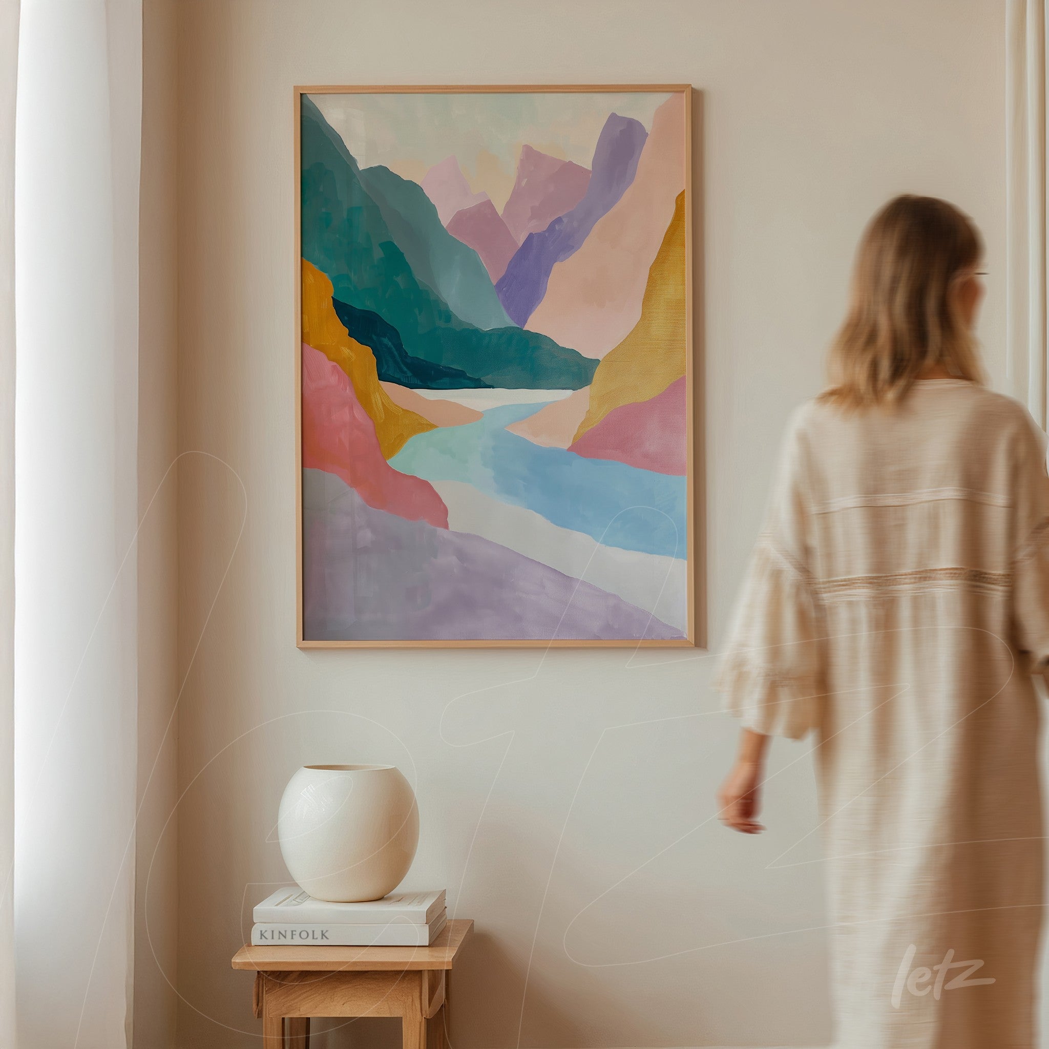 framed abstract landscape art piece featuring mountains and a river in soft colors, displayed on a neutral wall with a person blurred in the background