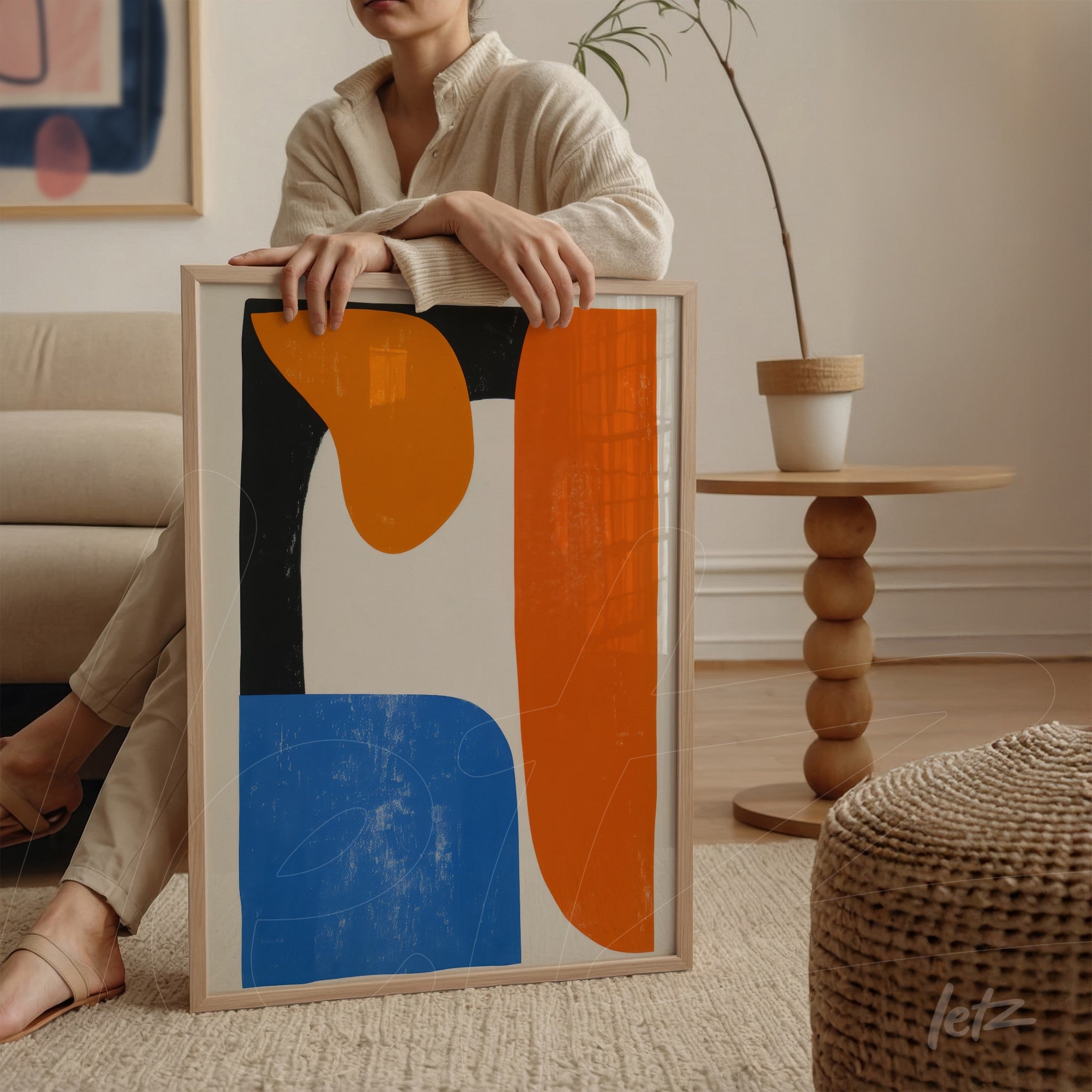 framed abstract art piece in shades of blue, orange and black held by a person sitting next to a sofa