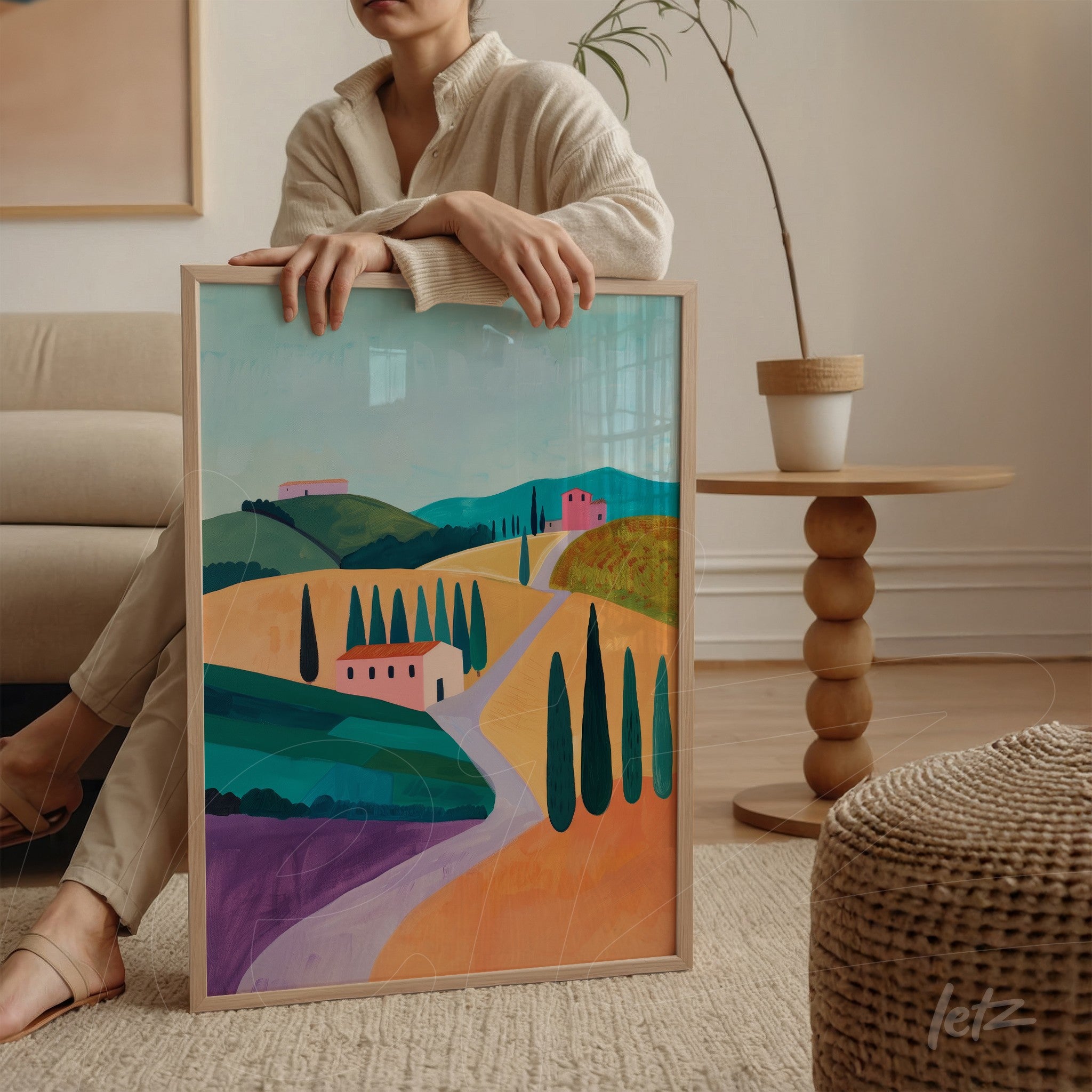 framed art piece featuring a vibrant landscape of hills, small villages, and cypress trees, held by a woman sitting in a modern interior