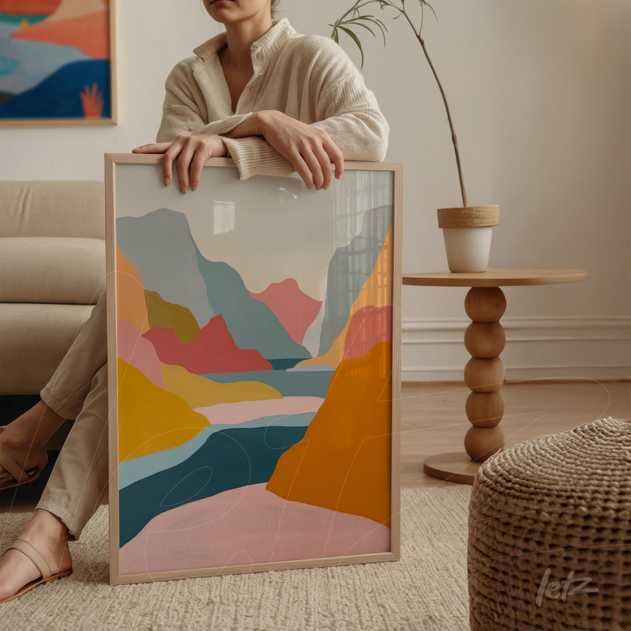 framed abstract art in shades of blue, pink, and orange held next to a woman beside a table
