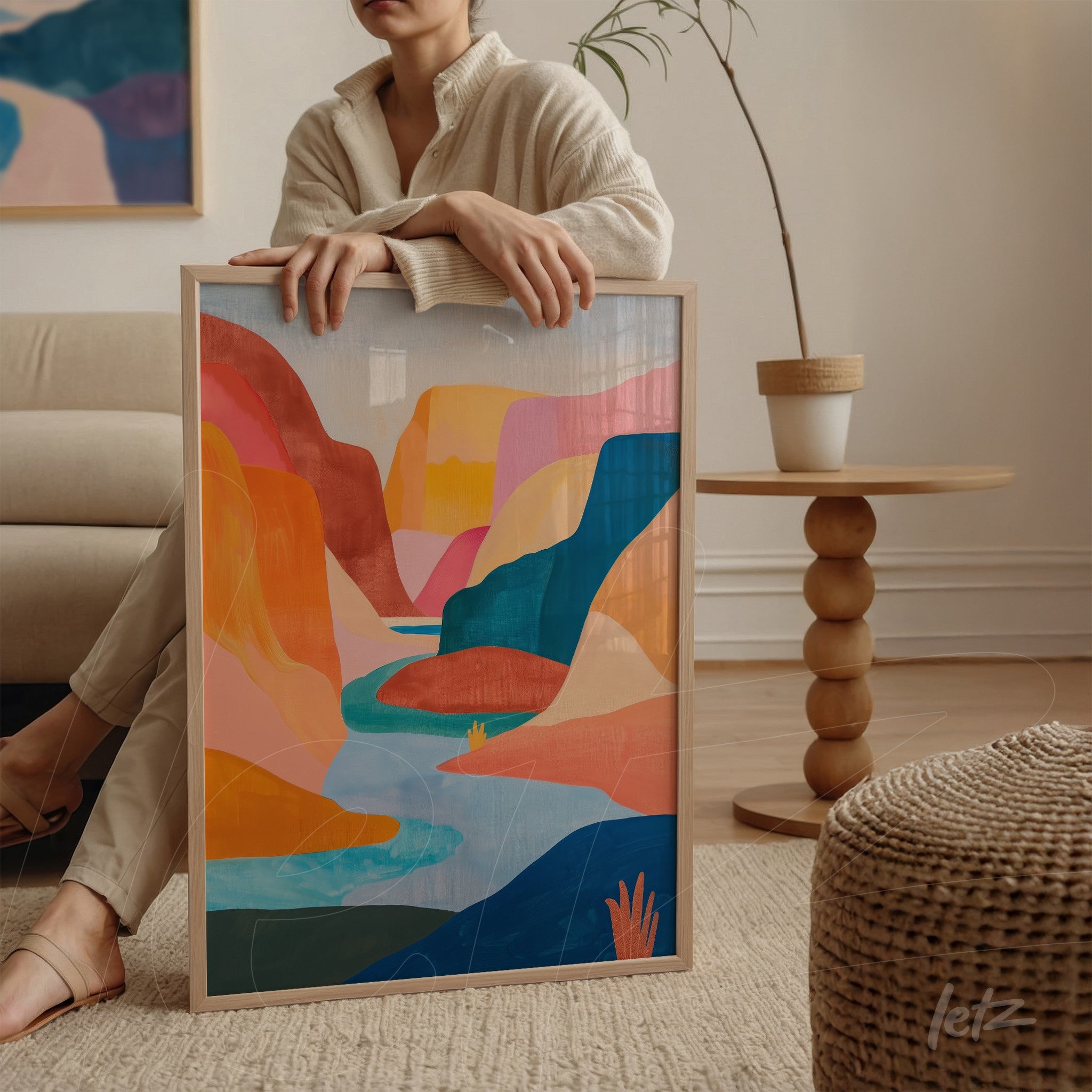 framed wall art featuring a vibrant abstract landscape in shades of orange, blue, and green, supported by a woman's leg in a naturally lit interior