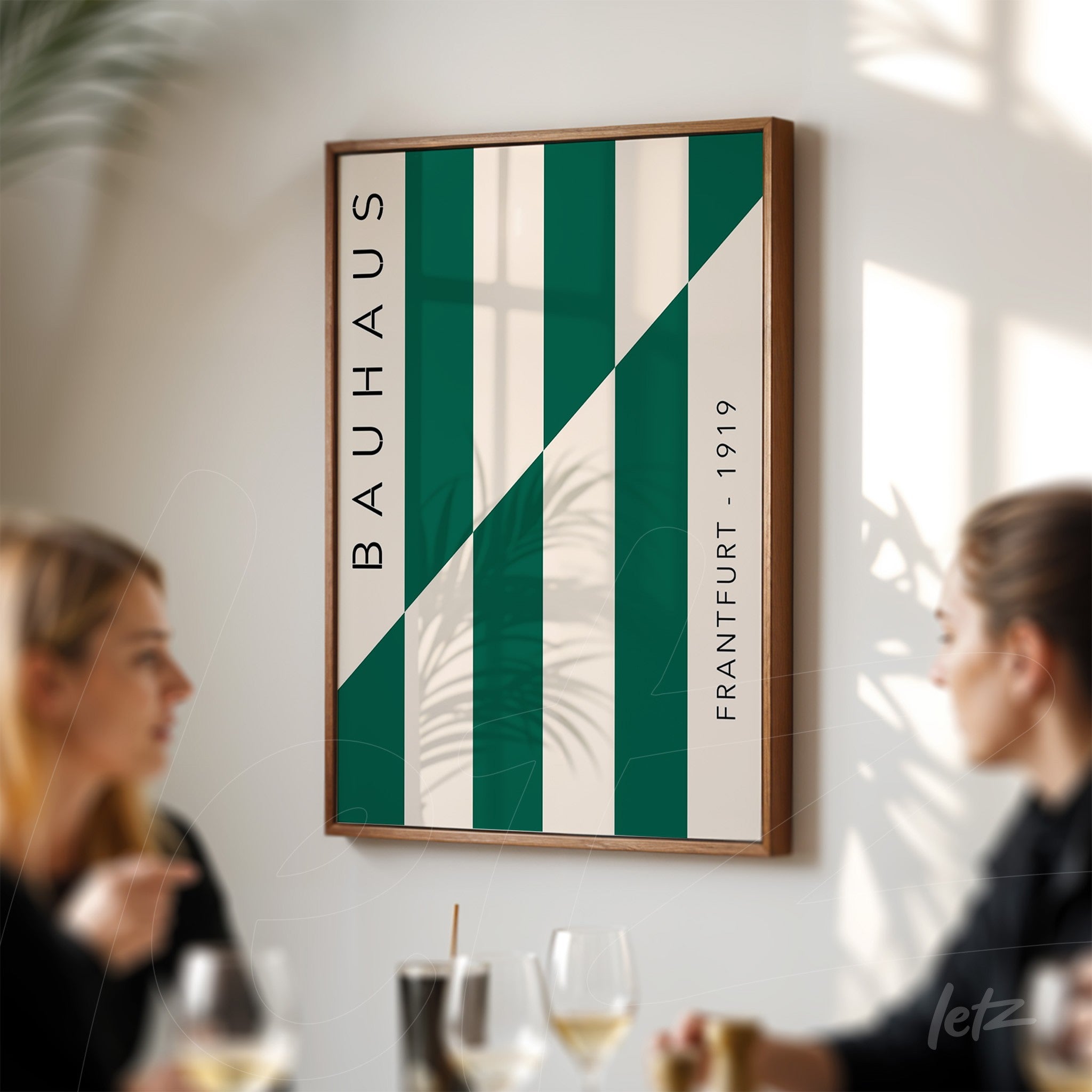 green and white geometric design canvas with brown wooden frame on a light wall, blurred figures of two people in the background