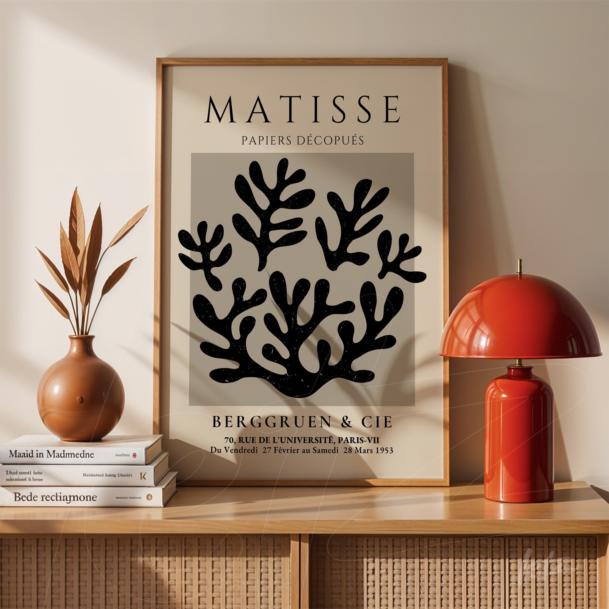 decorative wall art featuring leaf shapes in black on gray background, framed in light wood, displayed on a wooden shelf next to a ceramic vase and red lamp