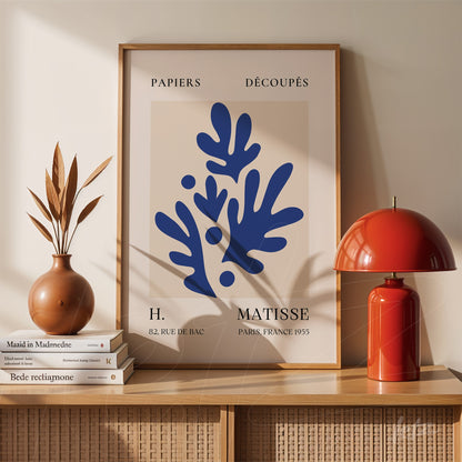framed abstract wall art in blue depicting organic shapes against a beige background displayed on a wooden shelf beside a red lamp and a vase