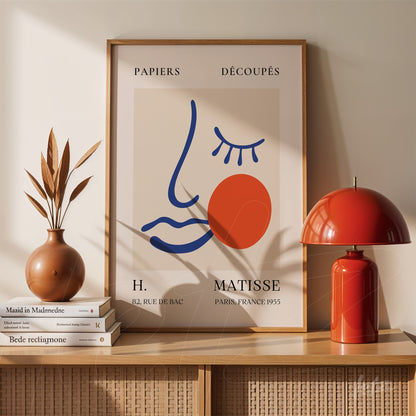 framed minimalist artwork featuring abstract face lines and a red circle, displayed on a table next to a red lamp