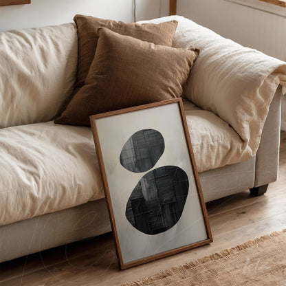 framed wall art featuring black and white abstract shapes with a light wood frame, leaning beside pillows on a beige sofa