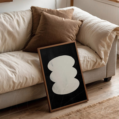 framed abstract wall art with black background and white shapes leaning against sofa with brown cushions