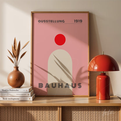 bauhaus art poster with red circle and pink background in light wood frame, displayed on wooden furniture beside a red lamp and dried leaves vase