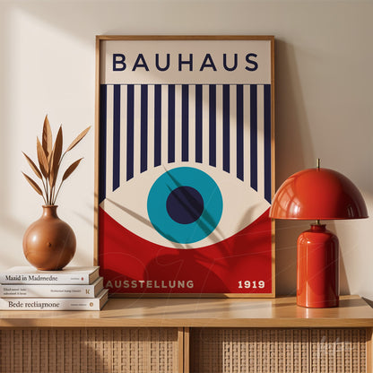 bauhaus style artwork with blue vertical stripes and a central eye, framed in light wood, displayed on a wooden cabinet with a red lamp and decorative vase nearby