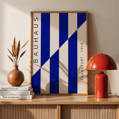 poster print with geometric art of blue and white stripes and the text 'bauhaus frankfurt 1919' in a light wood frame placed on light wooden furniture with a red lampshade and decorative vase