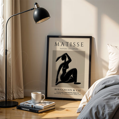 framed modern art depicting a female figure in black, leaning against a wooden stand next to a floor lamp
