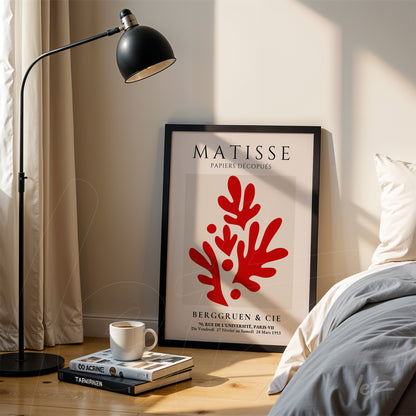 framed artwork by Matisse with coral red details against a light background, positioned beside a lamp and stacks of books