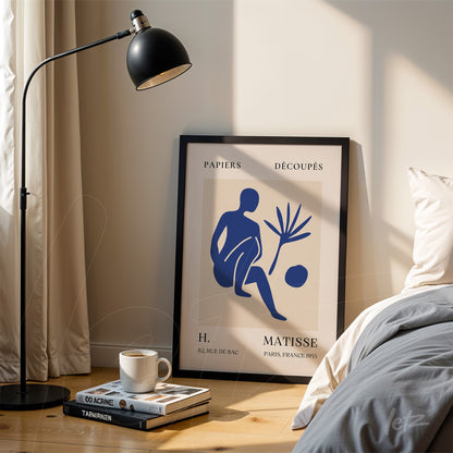 framed wall art featuring stylized blue human figures on a light background displayed next to a floor lamp and a stack of books on a wooden table