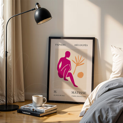 framed artwork by Henri Matisse featuring a stylized figure in shades of pink and orange, with botanical elements and a light background, displayed next to a lamp and coffee cup