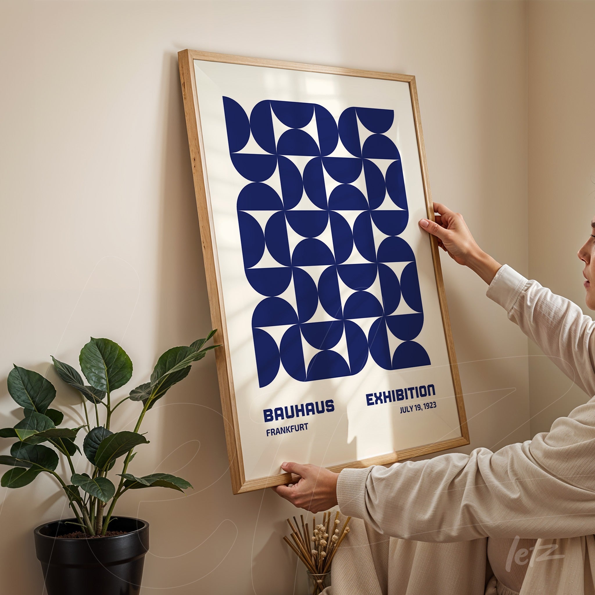poster with blue geometric design in light wood frame, being hung on a neutral wall next to a green plant