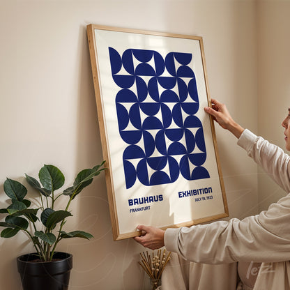 poster with blue geometric design in light wood frame, being hung on a neutral wall next to a green plant