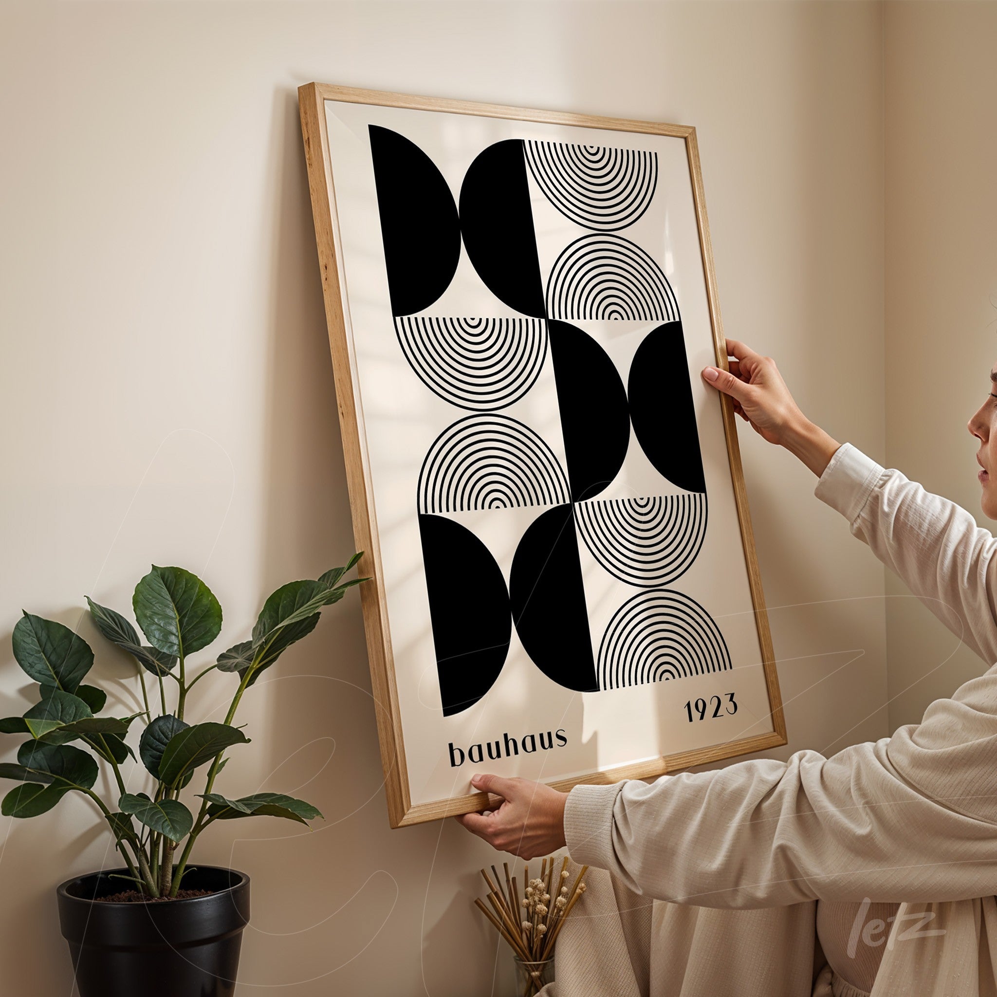 printed poster featuring geometric bauhaus style art in a light wooden frame being adjusted on a beige wall next to a black potted plant