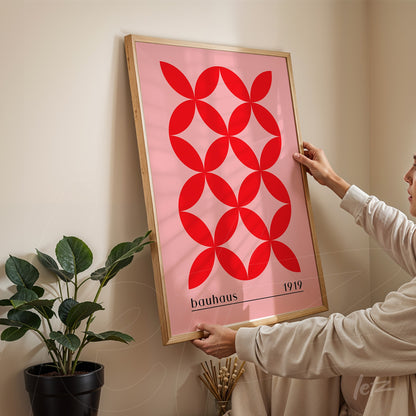 poster art with red geometric pattern on a pink background in a light wooden frame, displayed on a beige wall