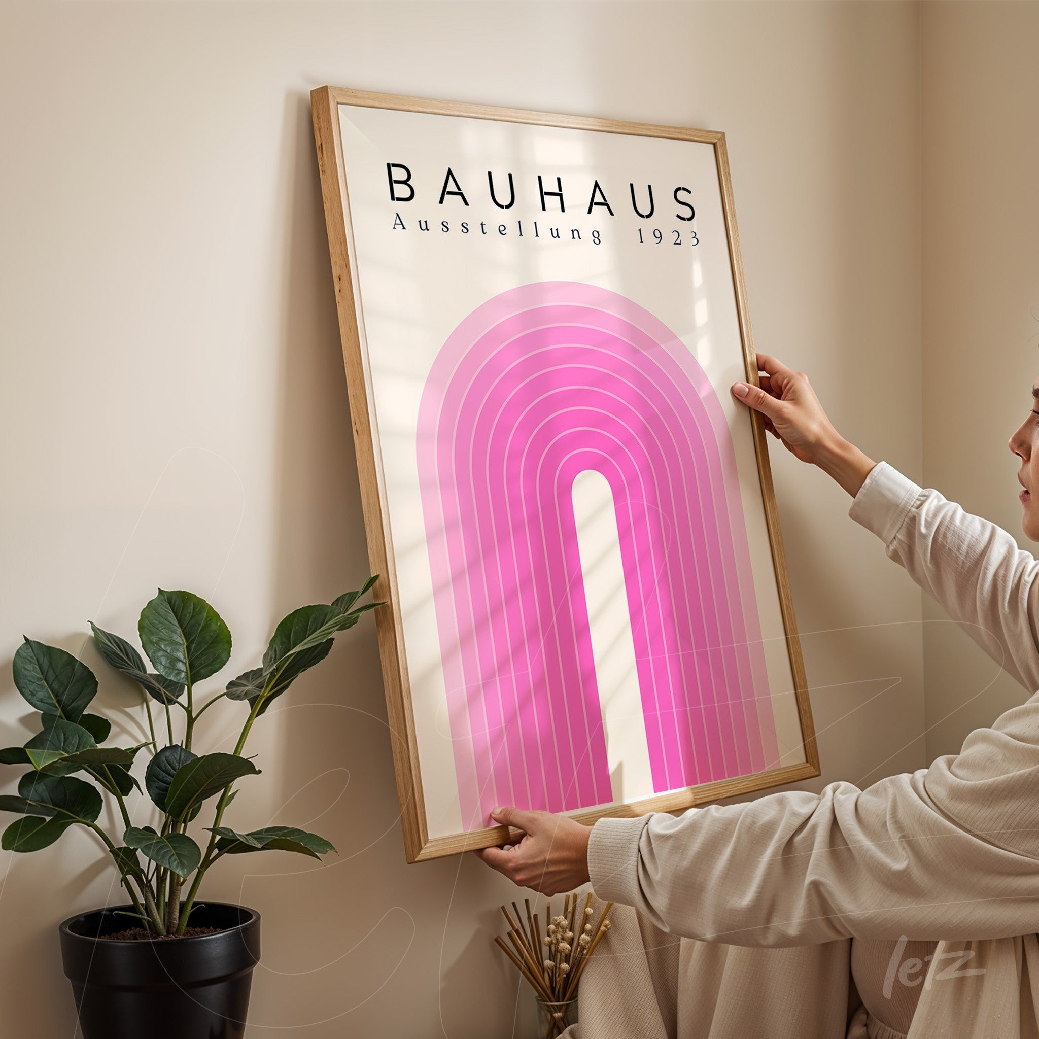 poster featuring a Bauhaus-style geometric design with a pink arch in a light wood frame being hung on a beige wall