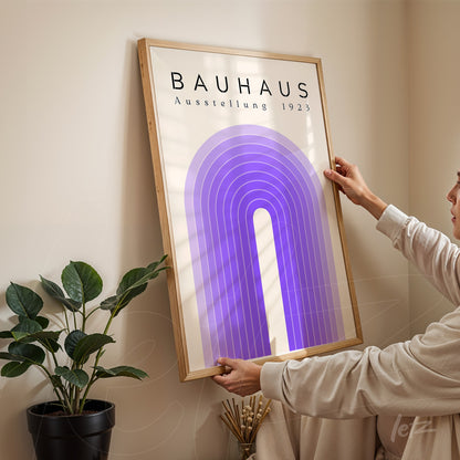 person adjusting bauhaus-style print with purple arch inside wooden frame on light wall beside a plant