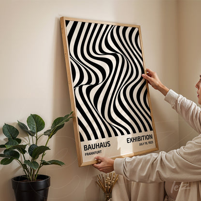 poster with black and white wavy line abstract pattern, framed in light wood, held by a person against a cream-colored wall