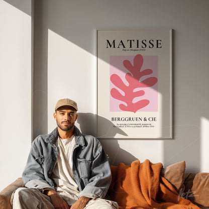 framed modern art piece featuring pink background and red leaves, displayed on a well-lit wall with a brown sofa and orange throw blanket