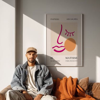 decorative framed art featuring a stylized face illustration by Henri Matisse, displayed on a well-lit wall next to a man sitting on a sofa