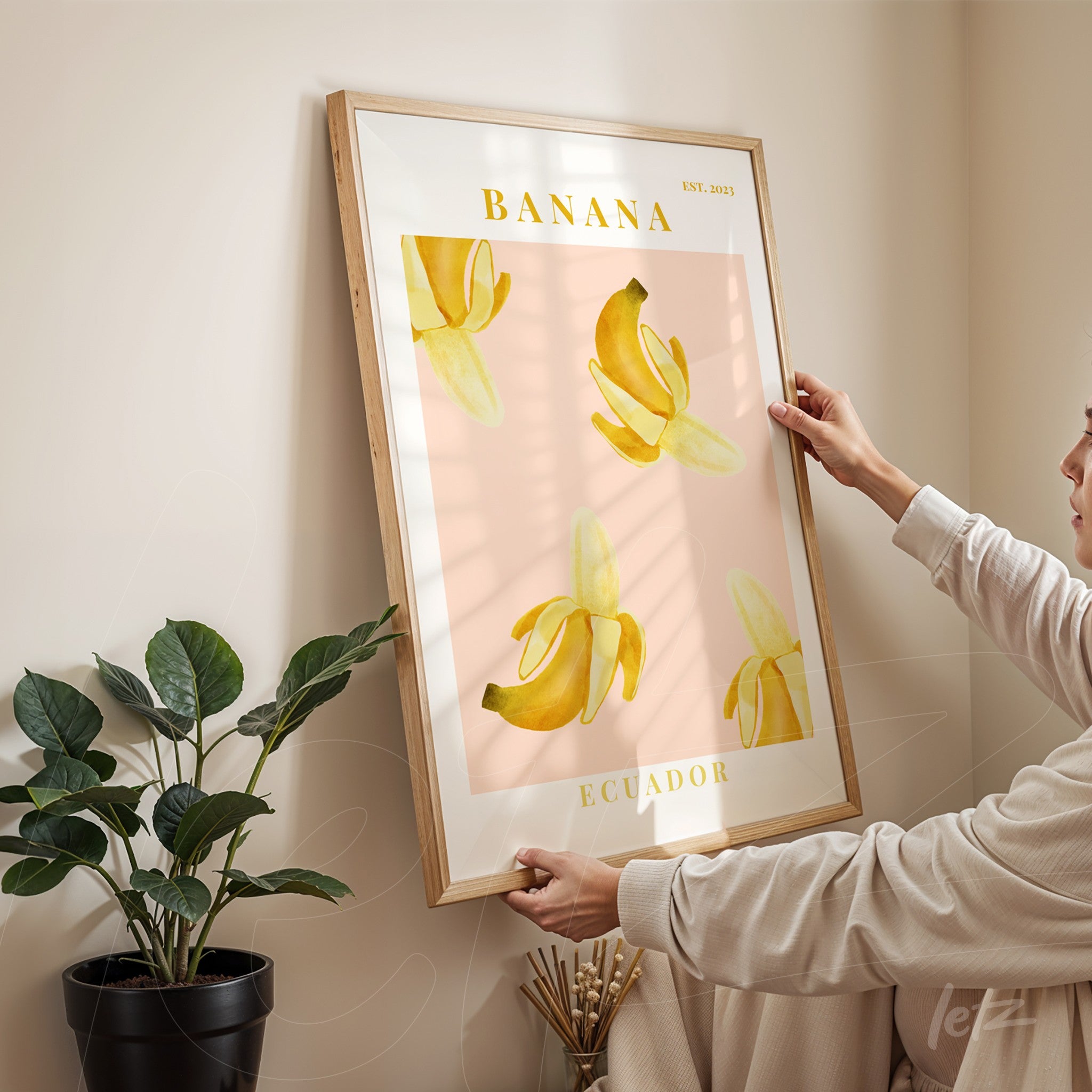 framed wall art featuring yellow bananas illustration on pink background held by a woman in front of a plant