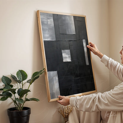 abstract wall art in shades of black and gray in a light wood frame held by a person in a neutral setting
