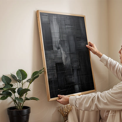 abstract wall art in black tones with a light wood frame being held by a person in a minimalist setting