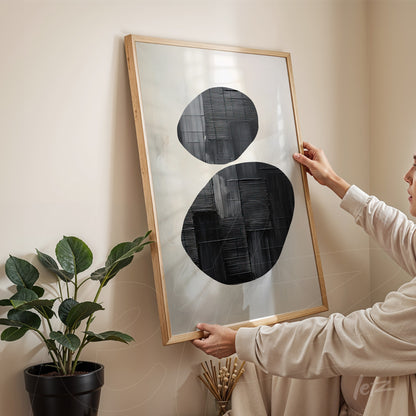 framed abstract black and white artwork held by a person in a cozy setting with a plant in the background