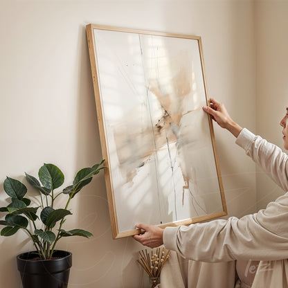 abstract artwork in neutral tones framed in light wood being held in an interior setting with a plant