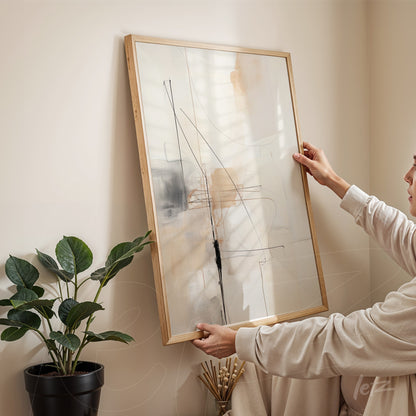 abstract wall art in light wood frame featuring subtle lines and shapes in neutral tones held by a person next to a green plant