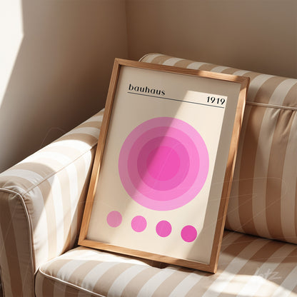 poster print of concentric pink circles in varying shades with 'bauhaus 1919' text displayed in wooden frame on striped sofa