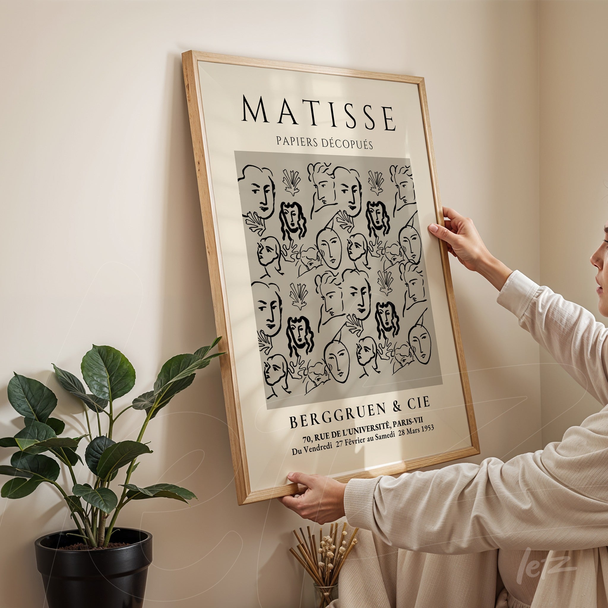 decorative wall art featuring a black and white illustration of minimalistic faces, light wooden frame, displayed on wall with a plant beside it