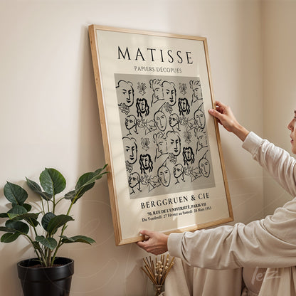 decorative wall art featuring a black and white illustration of minimalistic faces, light wooden frame, displayed on wall with a plant beside it