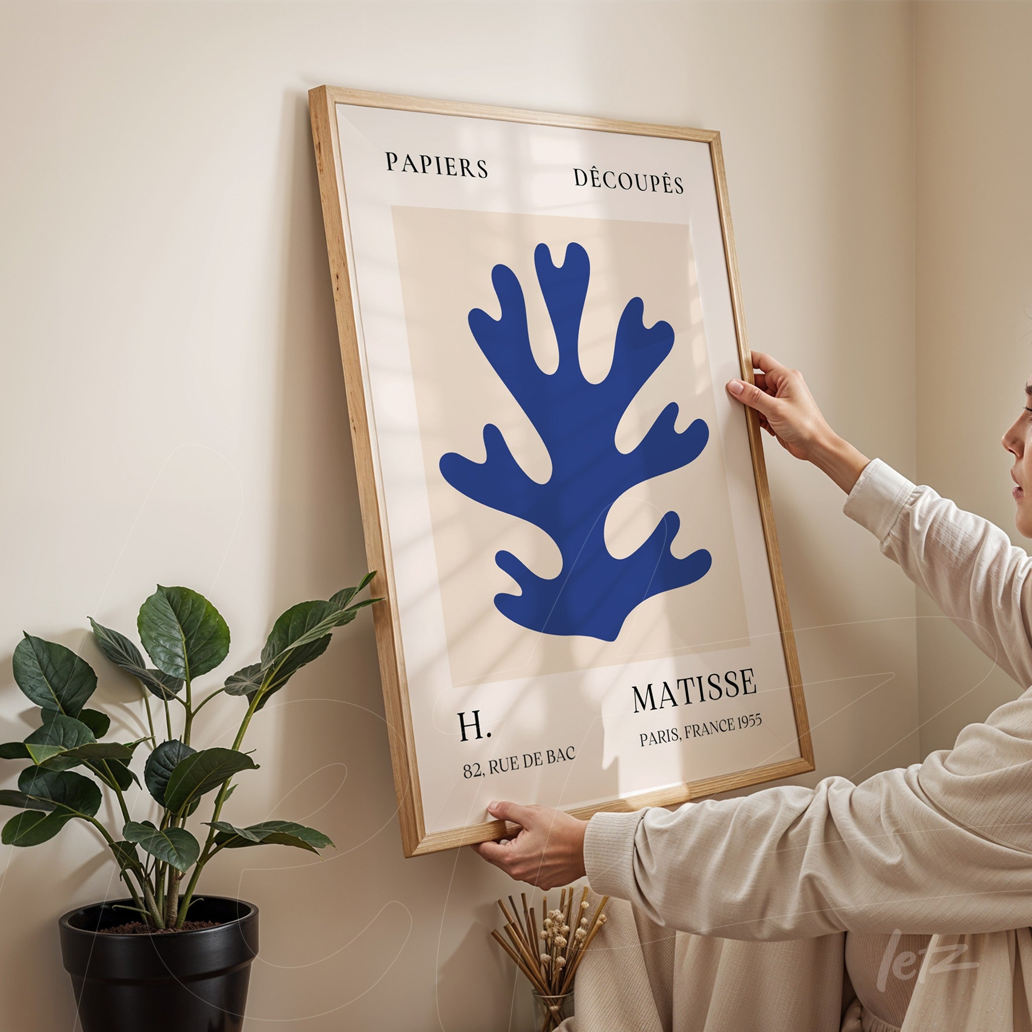 framed modern art piece featuring a blue coral shape in light wood frame held by a person in a room with a plant